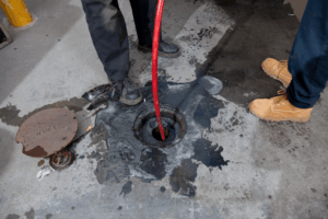 Two people stand near an uncovered manhole as a red hose is inserted for hydro jetting solutions Philadelphia; a manhole cover labeled "ELEVATOR" lies on the ground nearby.